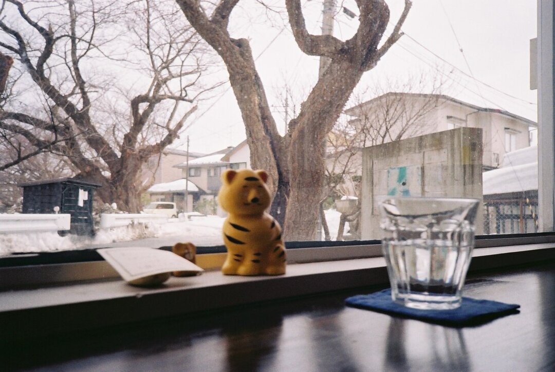A film photo of a small yellow tiger figurine in the window of a cafe. Snowy street and wintered trees can be seen in the background, a glass of water in the foreground